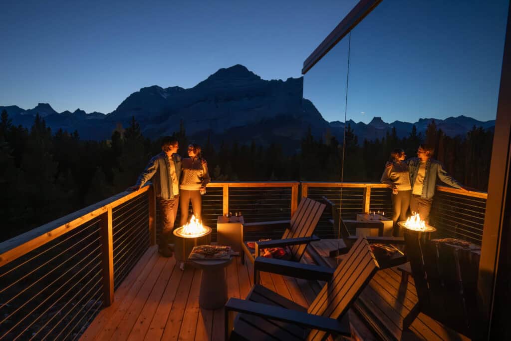 A couple enjoying the scenic mountain view from a wooden deck with fire pits at dusk, surrounded by nature and mountains.