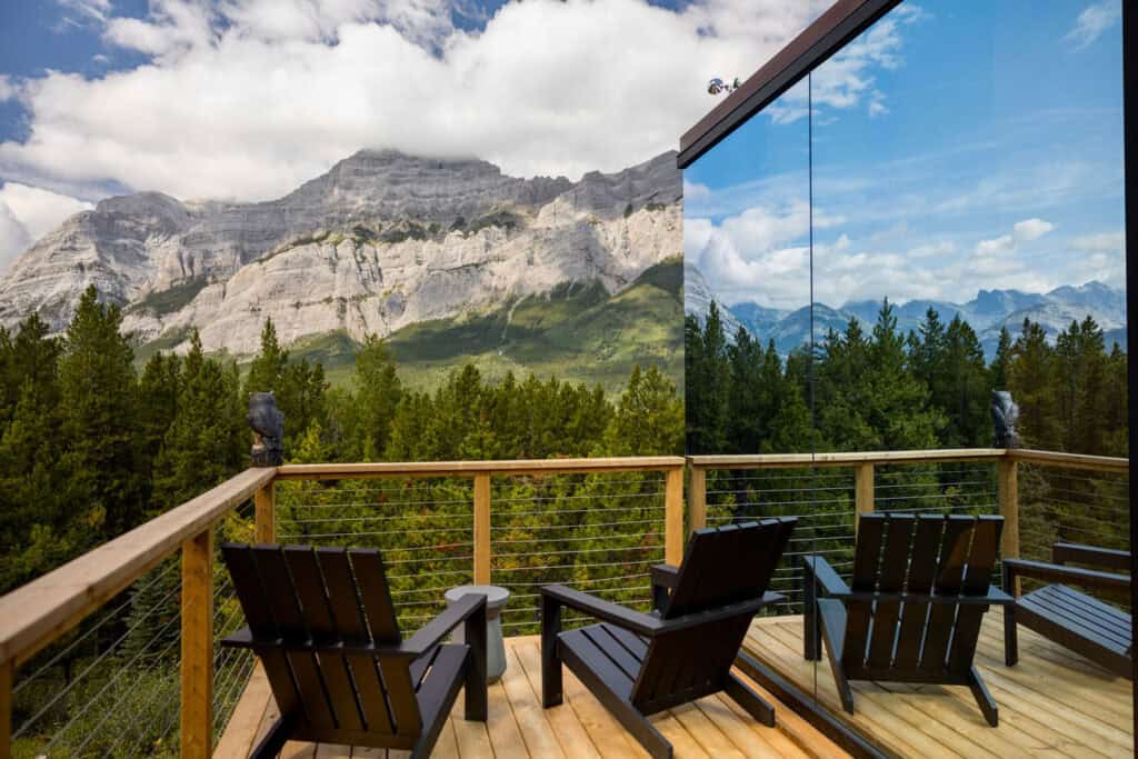 Scenic mountain view from a wooden balcony with chairs, overlooking lush forests and rugged peaks, blending natural beauty with contemporary architecture.