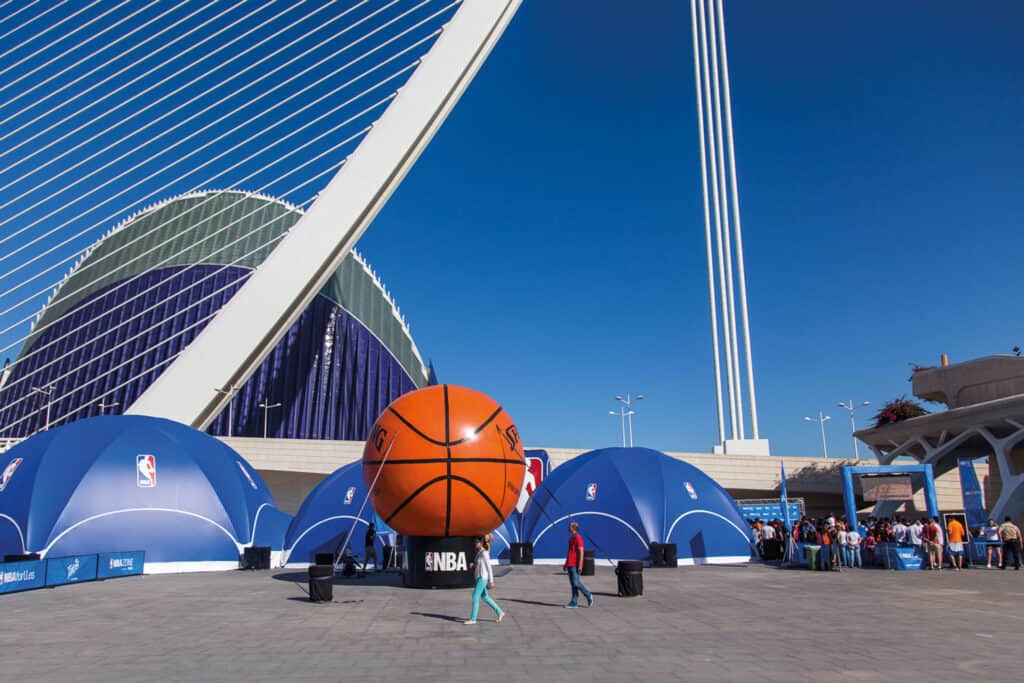 Modern sports venue featuring a large basketball sculpture, innovative design, and surrounding tents, highlighting the integration of sports and entertainment industries.
