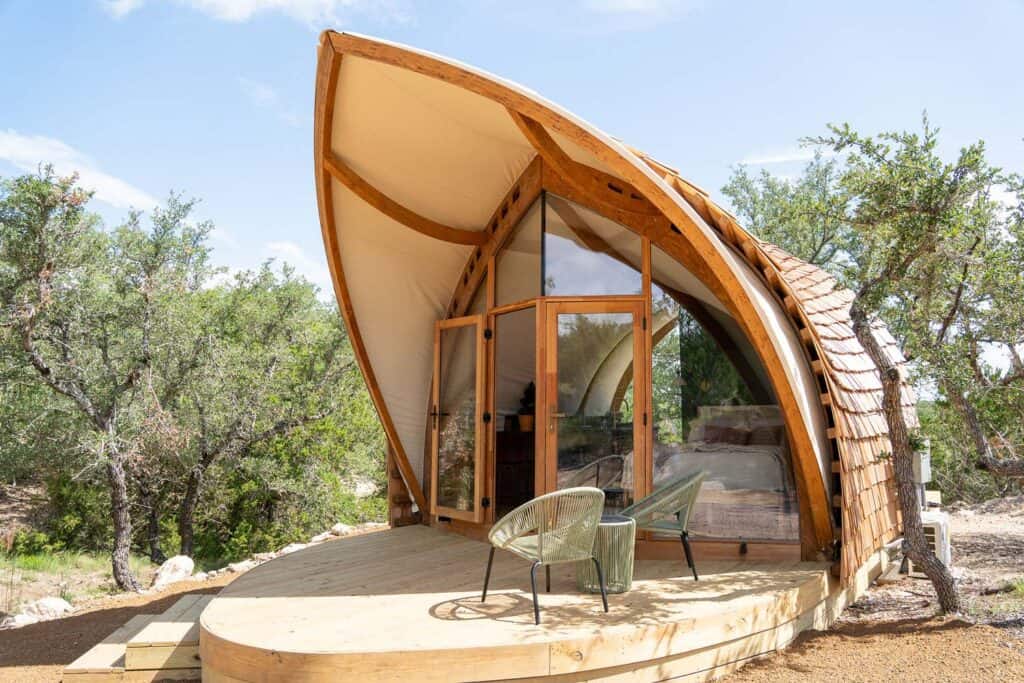 A modern tiny house featuring a unique curved wooden structure and large glass windows, set in a natural outdoor environment with trees and clear skies.