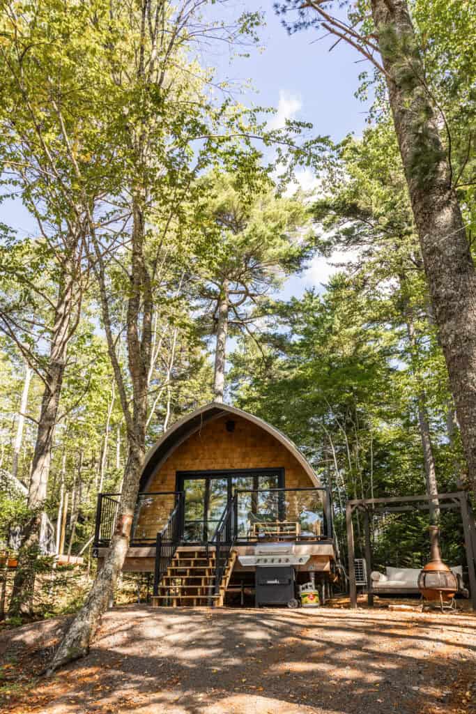 A-Frame cabin nestled among lush green trees in a forest setting, featuring a wooden deck with outdoor seating and a barbecue grill, perfect for nature retreats.