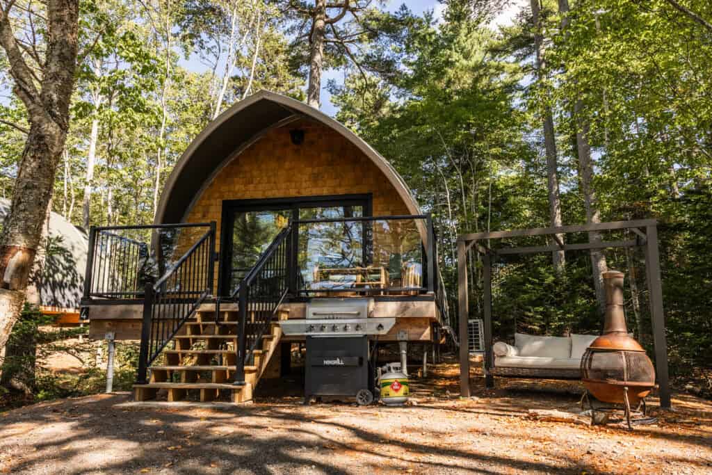 A-Frame cabin nestled among tall trees in a lush forest, featuring a wooden deck with outdoor seating and a fire pit, perfect for nature retreats and outdoor relaxation.