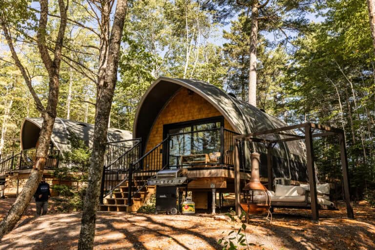 A-Frame cabin nestled among tall trees in a forest setting, featuring a wooden deck, outdoor seating, and a barbecue grill, perfect for nature retreats.