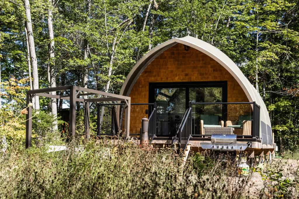 A contemporary cabin with a curved roof nestled among lush green trees, featuring a small deck with outdoor seating and glass doors opening to nature.